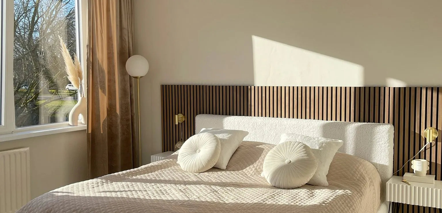 Solbeskinnet moderne soveværelse med en beige quiltet seng, runde puder, Akupanel-lameller som hovedgærde og minimal indretning ved siden af et stort vindue med gennemsigtige gardiner.